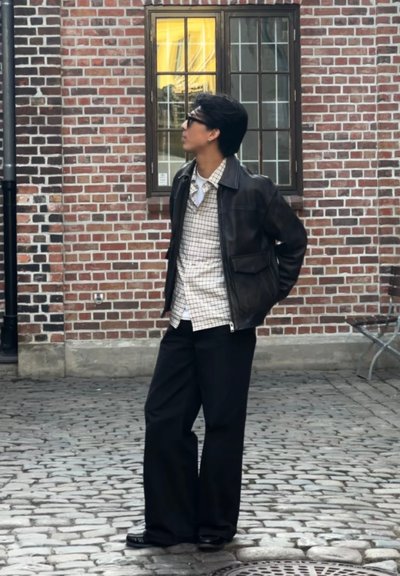 Chaqueta de cuero marrón sobre una camisa a cuadros blanca y beige, pantalones de pierna ancha negros y zapatos negros, de pie en una calle empedrada.