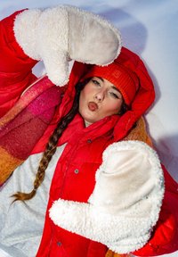 Γυναίκα με κόκκινο χειμερινό παλτό και καπέλο, ξαπλωμένη στο χιόνι, σχηματίζει φιλί με τα χέρια της που φοράνε λευκά γάντια και απλώνονται προς την κάμερα.