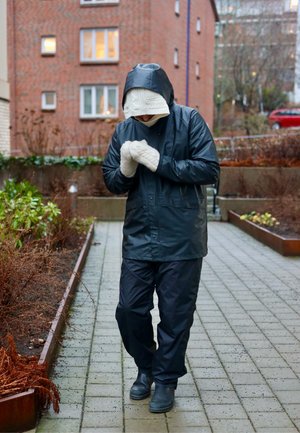 Chaqueta y pantalones impermeables negros, guantes tejidos blancos con textura y botas negras. Entorno al aire libre con plantas y edificios de ladrillo.