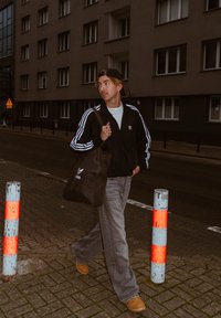 Melnā Adidas jaka ar baltām svītrām, gaiši zils krekls, pelēkas džinsu bikses, brūni zābaki, nēsājot brūnu tote somu, pastaigājas pie urbānām ēkām.