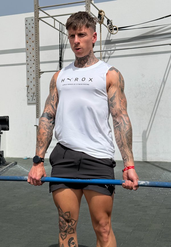 Hombre con tatuajes levantando una barra con pesas al aire libre en una zona de fitness, vistiendo una camiseta blanca sin mangas y pantalones cortos negros.