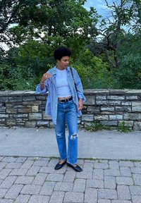 Kék és fehér csíkos, oversized gombos ing, fehér, testhezálló póló, kopott kék farmernadrág és fekete slip-on cipők egy kőfal előtt.