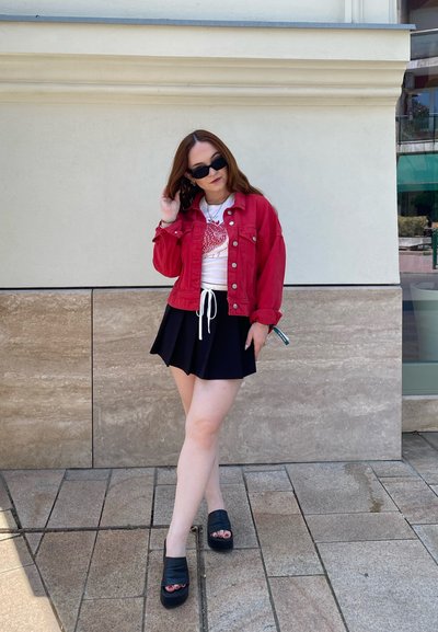 Chaqueta de mezclilla roja sobre una camiseta blanca con un gráfico, combinada con una falda corta de pliegues negra y sandalias de plataforma negras, posando al aire libre.