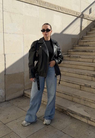 Mujer con chaqueta de cuero negra, top negro, vaqueros de pierna ancha azul claro, zapatillas blancas y gafas de sol negras, de pie cerca de unas escaleras de piedra.
