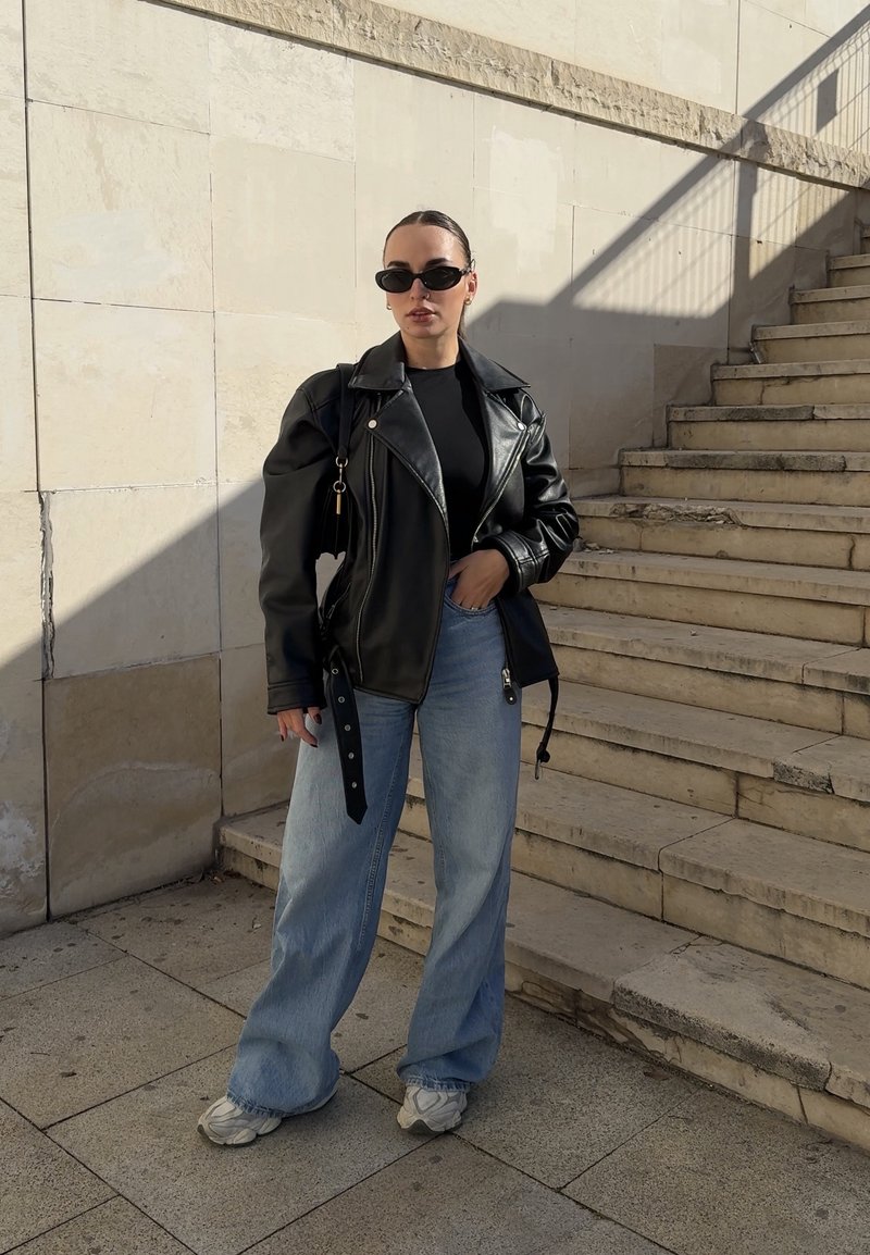 Donna con giacca di pelle nera, top nero, jeans a gamba larga azzurri chiaro, sneakers bianche e occhiali da sole neri in piedi vicino a una scalinata di pietra.