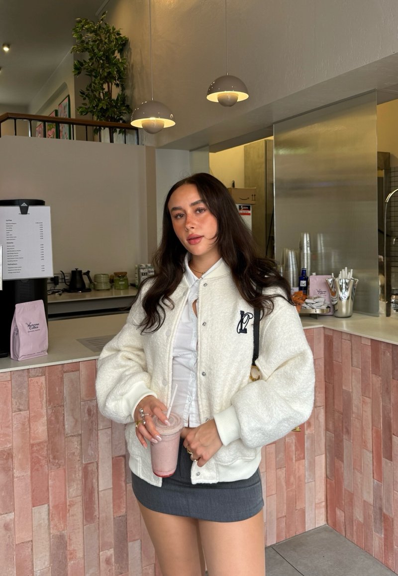 Veste blanche, duveteuse et surdimensionnée avec des poignets côtelés, portée sur une chemise blanche à boutons et une jupe grise. Smoothie rose clair à la main.