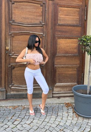 Top corto de textura rosa, leggings capri blancos y chanclas blancas. De pie junto a una puerta de madera y una planta en maceta sobre una superficie de adoquines.