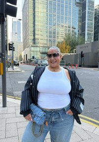 Casaco oversized às riscas pretas e brancas, top branco, mala azul clara com corrente, jeans de ganga largos; ambiente urbano com edifícios modernos.
