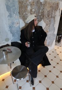 Femme portant un manteau noir et un béret crème, assise les jambes croisées sur un banc dans un café, tenant une tasse en métal, regardant sur le côté.