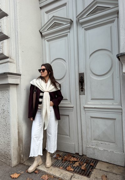 Mujer con gafas de sol, chaqueta oscura, bufanda blanca, pantalones blancos y botas beige, de pie frente a unas puertas dobles ornamentadas blancas en una acera cubierta de hojas.