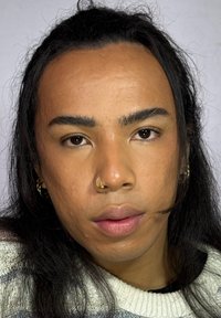 Close-up van een persoon met donker, lang haar en een neusring, die een lichtgekleurde trui met horizontale strepen draagt. Heeft een gladde huid en neutrale make-up.