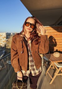 Giacca in camoscio marrone con due tasche frontali e dettagli blu, indossata sopra una camicia a quadri. Grandi occhiali da sole scuri e pantaloni marroni. Borsa con pelliccia.