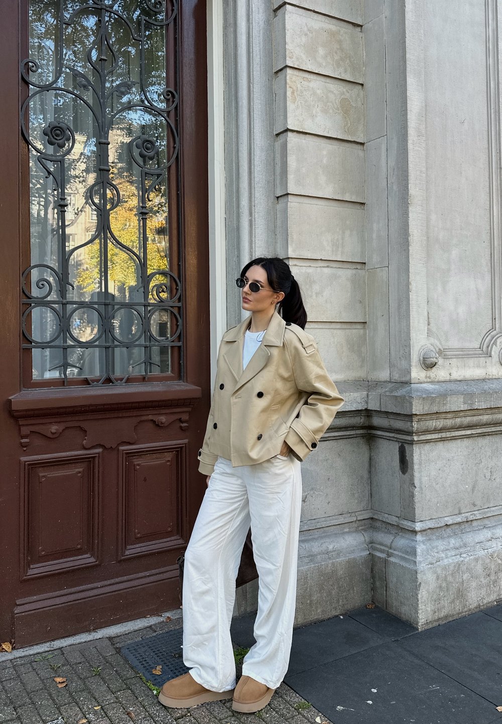 Béžový dvojradový kabát, biele nohavice s širokými nohavicami a hnedé obuv. Okrúhle slnečné okuliare dopĺňajú outfit pred textúrovanou stenou.