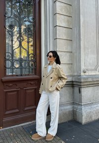 Béžový dvojradový kabát, biele nohavice s širokými nohavicami a hnedé obuv. Okrúhle slnečné okuliare dopĺňajú outfit pred textúrovanou stenou.