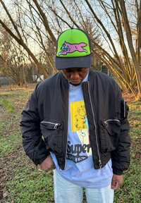 Fekete bombázójáték dzseki cipzáras zsebekkel, világoskék grafikás póló nyomattal, valamint zöld-fekete sapka rajzfilm mintával.