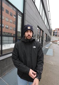 Homme portant une veste noire North Face et un bonnet Carhartt bleu marine ajustant sa manche à l'extérieur près d'un bâtiment moderne par une journée nuageuse.