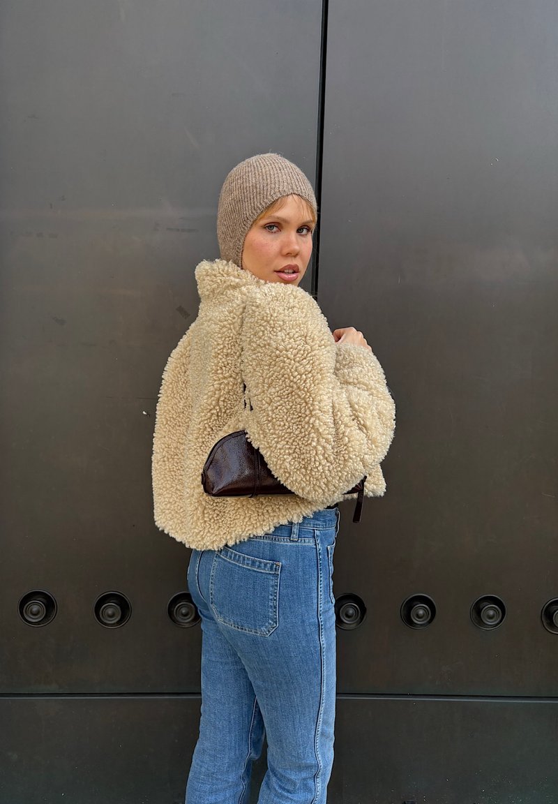 Veste en polaire beige avec une texture, associée à un jean en denim bleu clair. Sac à main marron foncé de forme courbée, porté à l'épaule.