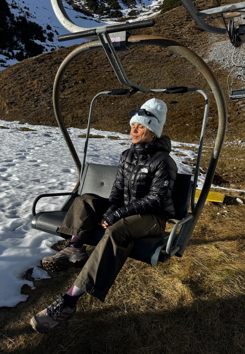Femme portant un chapeau blanc et une veste noire assise sur un télésiège avec en arrière-plan des pentes de montagne enneigées et herbeuses.