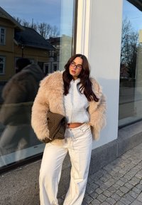 Beżowy płaszcz ze sztucznego futra z szerokimi rękawami, szara krótka bluza z kapturem i białe spodnie o szerokich nogawkach. Trzyma brązową, fakturowaną torebkę. Tło z lustrzaną powierzchnią.