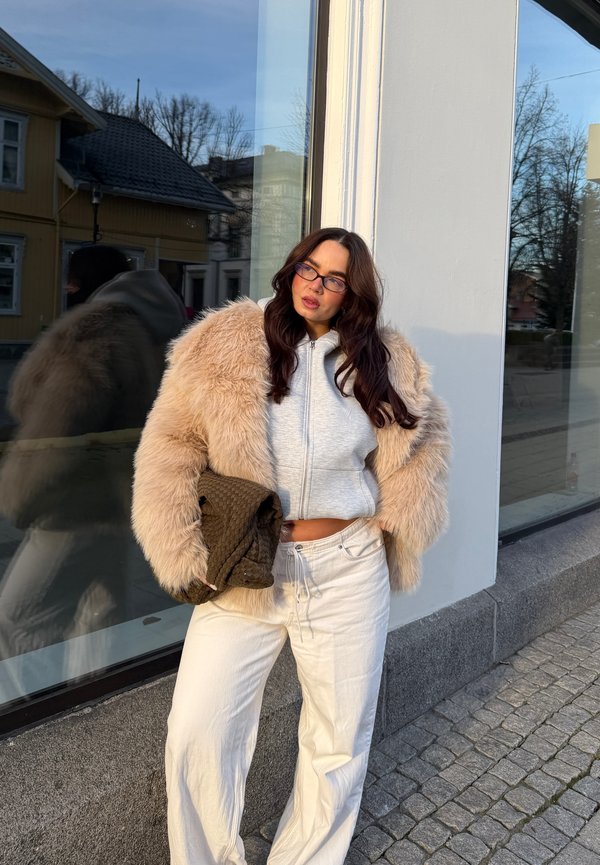 Beige fuskpälsjacka med vida ärmar, grå kort hoodie och vita vida byxor. Håller en brun texturerad handväska. Reflekterande yta i bakgrunden.
