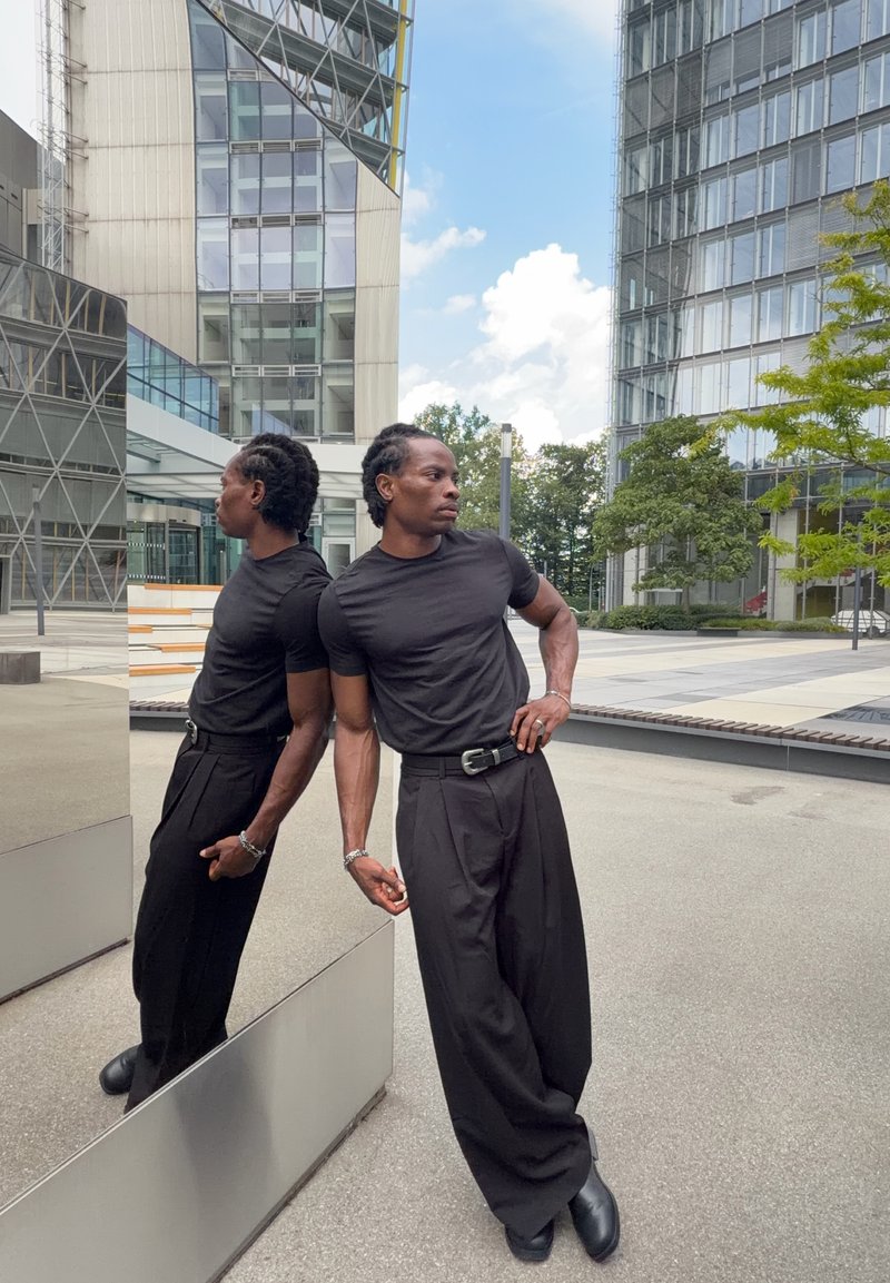 Homme en chemise et pantalon noirs appuyé contre un mur réfléchissant sur une place urbaine avec des bâtiments modernes en verre et des arbres en arrière-plan.