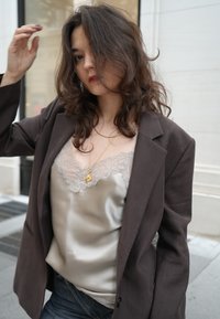 Barna túlméretezett blézer szatén csipkedíszítéses kombiné fölött, farmerrel párosítva. Puha hullámok keretezik az arcot, arany nyaklánc díszítéssel.