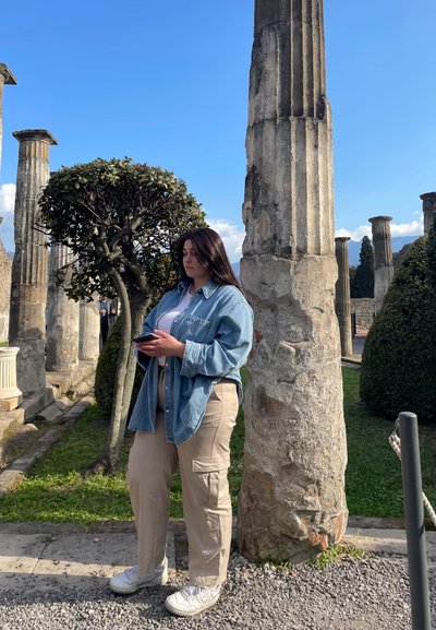 Una persona está de pie junto a una columna de piedra desgastada, vistiendo una camisa de mezclilla azul clara y pantalones cargo beiges, sosteniendo un teléfono inteligente.