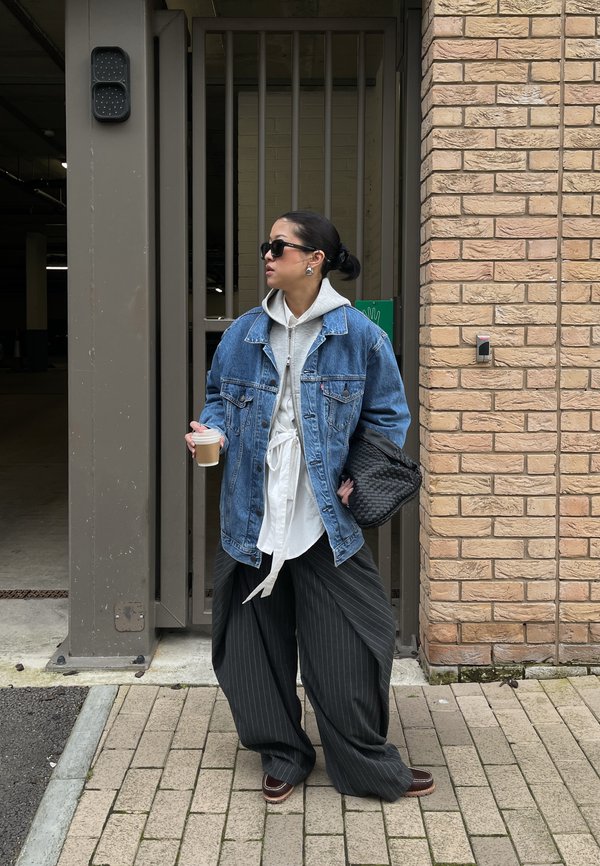 Person wearing oversized denim jacket, white hoodie, pinstripe wide pants, sunglasses, holding coffee cup and black woven bag by brick wall.