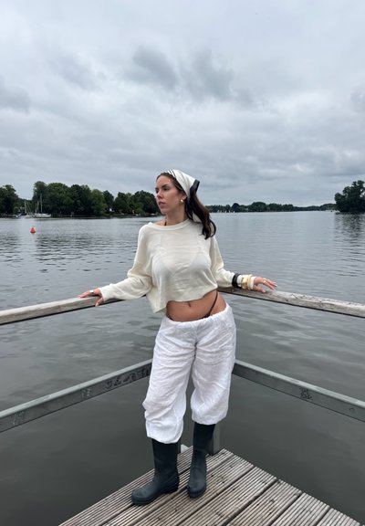 Suéter blanco corto, pantalones blancos holgados y botas de goma negras. Cabello recogido con una diadema en blanco y negro, de pie junto al agua. Cielo nublado.