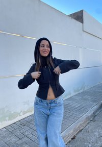 Črna zip-up kapuca s prilagojenim dizajnom, rebrastimi manšetami in kapuco, v kombinaciji s svetlo modrimi visokimi širokima hlačami.