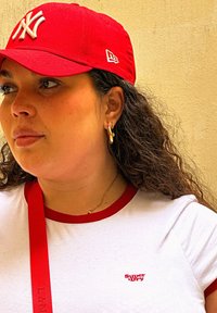 Gorra roja de los New York Yankees, camiseta blanca con ribete rojo y pequeño logo, pendientes de aro dorados y una correa roja cruzada sobre el hombro. Cabello rizado.