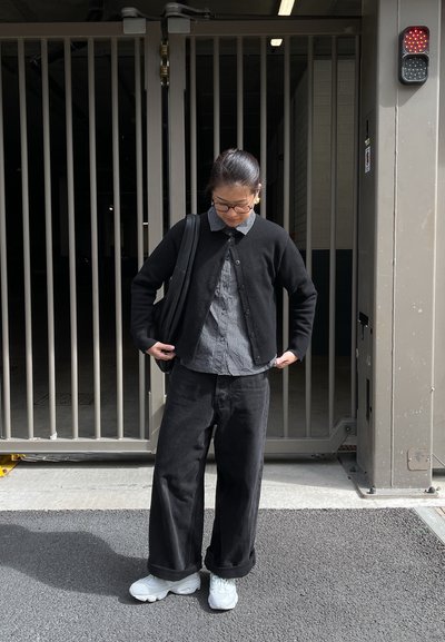 Cárdigan negro de punto sobre una camisa de cuadros abotonada, combinado con pantalones anchos negros y zapatillas blancas gruesas. El fondo presenta una puerta.