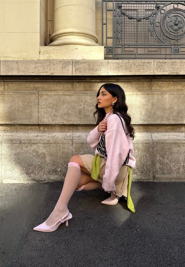 Une femme en pull rose, jupe beige et chaussures à talons roses pose assise contre un bâtiment en pierre doté d'une rampe en métal ornée.