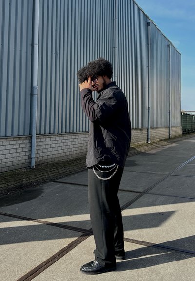 Camisa negra de manga larga, pantalones negros holgados con detalle de cadena y zapatos negros. Frente a un fondo de paneles grises verticales. Luz del día brillante.