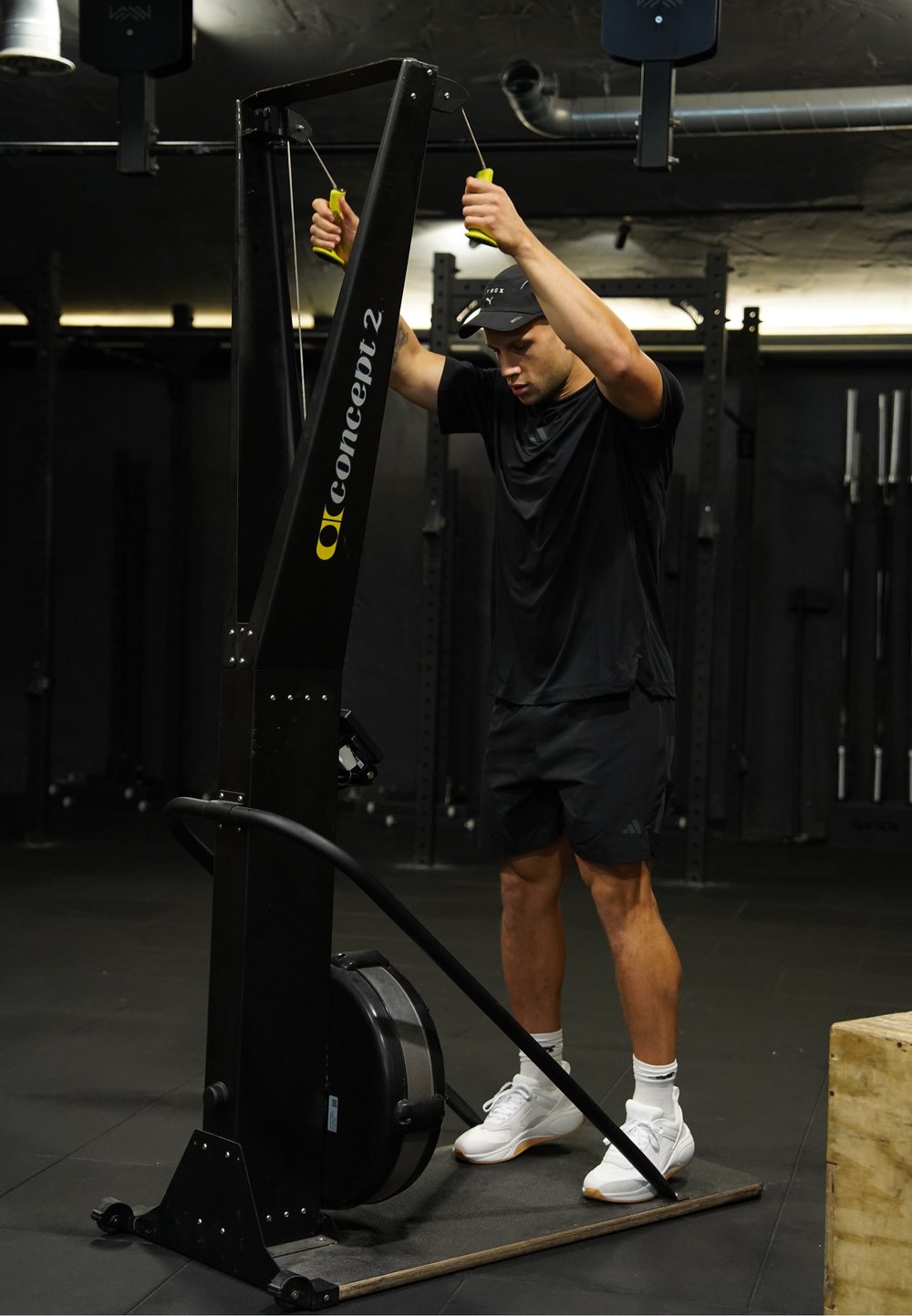 Άνδρας με μαύρα ρούχα γυμναστικής χρησιμοποιεί μηχάνημα Concept2 ski erg σε χαμηλά φωτισμένο γυμναστήριο με ράφια βαρών στο παρασκήνιο.