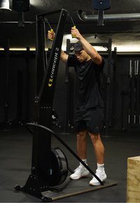 Άνδρας με μαύρα ρούχα γυμναστικής χρησιμοποιεί μηχάνημα Concept2 ski erg σε χαμηλά φωτισμένο γυμναστήριο με ράφια βαρών στο παρασκήνιο.