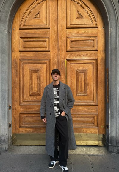 Abrigo de lana gris, camiseta gráfica negra con texto, pantalones negros y zapatillas, apoyados contra una puerta de madera con paneles detallados.