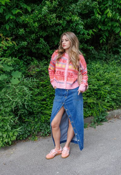 Cárdigan rosa con estampado, falda de mezclilla azul con un slit delantero y botones, combinada con zapatillas deportivas rosa claro. Fondo de follaje verde.