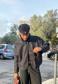 Uomo con giacca e pantaloni neri che sistema la manica all'aperto vicino a auto parcheggiate e alberi, tenendo una borsa in pelle marrone nella mano sinistra.