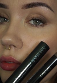 Close-up van een gezicht met een lichte huid, een gouden neusring en nude lippenstift. Twee donkergroene cosmeticaproducten worden dichtbij het gezicht vastgehouden.