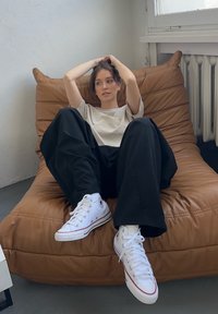 Bean bag chair in soft brown leather, model wearing a light beige shirt and loose black pants, featuring white sneakers with red accents.