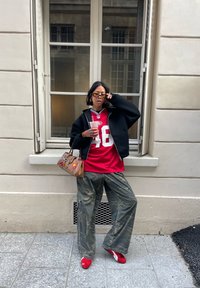 Rød fotballtrøye med hvitt nummer 48, svart hettegenser med glidelås, kamuflasje vide bukser, røde sko og brun håndveske med dekorative detaljer.