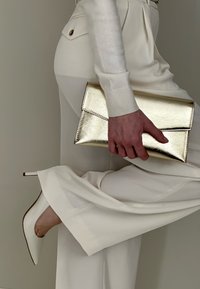 Femme en tenue crème tenant une pochette dorée brillante, levant une jambe pour montrer une chaussure à talon aiguilles pointue de couleur crème assortie.
