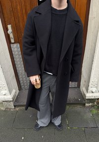 Cappotto di lana nero sopra un maglione di lana nero, abbinato a pantaloni a righe grigie. Anelli d'argento adornano una mano, che tiene una tazza di carta.