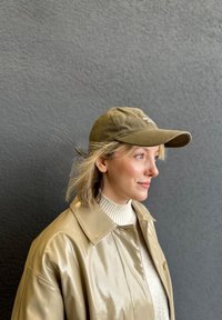 Olivengrønn bomullslue med buet skjerm, båret med en glansfull beige frakk og en kremfarget genser, mot en teksturert grå bakgrunn.