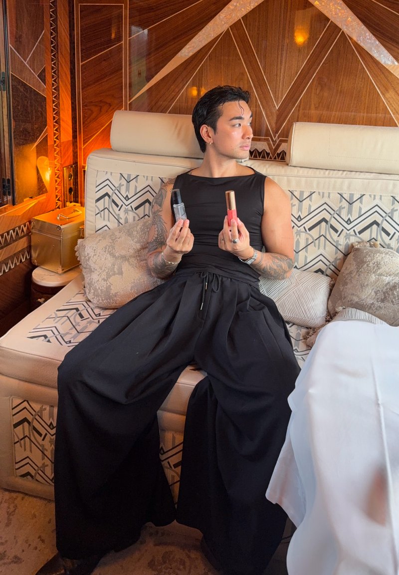 Personne tatouée assise sur un canapé à motifs, tenant deux produits de maquillage, regardant de côté dans une pièce chaleureusement éclairée avec des panneaux en bois.