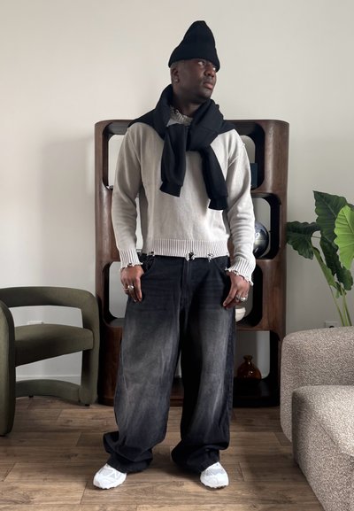 Hombre con gorro negro, suéter blanco desgastado, pantalones negros oversize y zapatillas blancas, de pie en una habitación con suelo de madera y plantas.