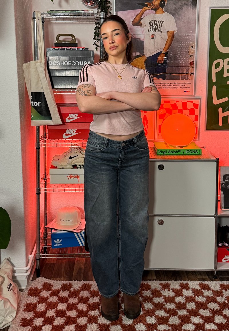 Jeune femme aux bras croisés, portant un t-shirt rose Adidas et un jean, se tient dans une pièce avec des boîtes à chaussures, des affiches et une lampe rouge-orangée.