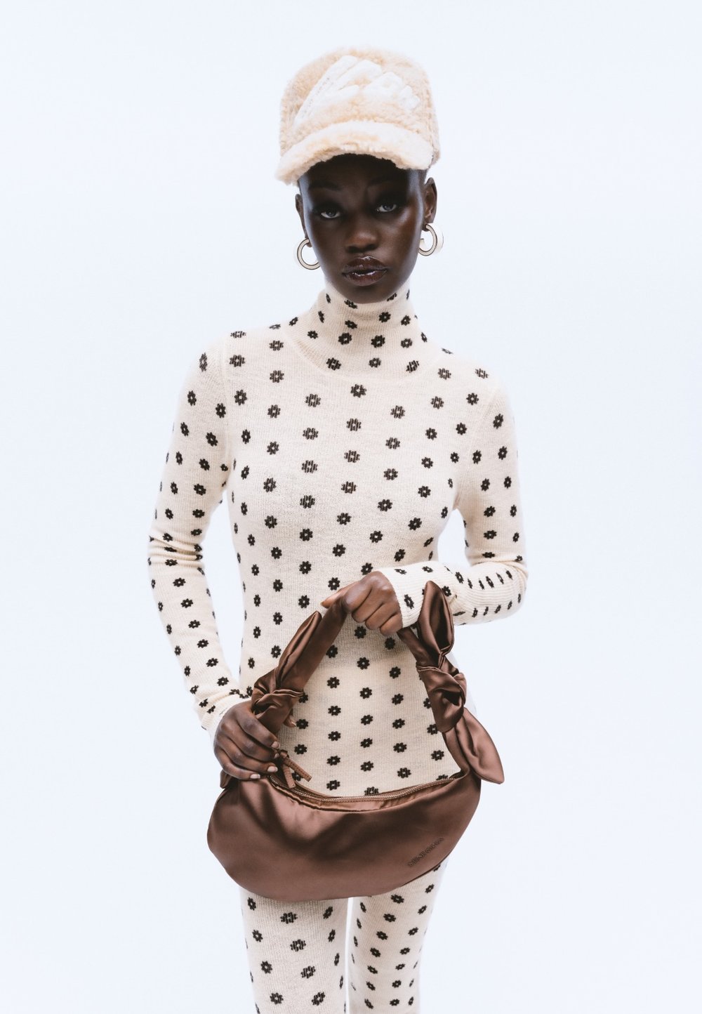 Outfit in crema con dolcevita a maglia con motivo floreale nero; tiene in mano una borsa in satin marrone con due manici annodati. Cappello di pelliccia beige.