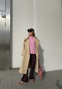 Beige trenchcoat, rosa genser, mørke treningsbukser med hvite striper, brun caps, mørke solbriller og brune sko. Rosa veske på skulderen.